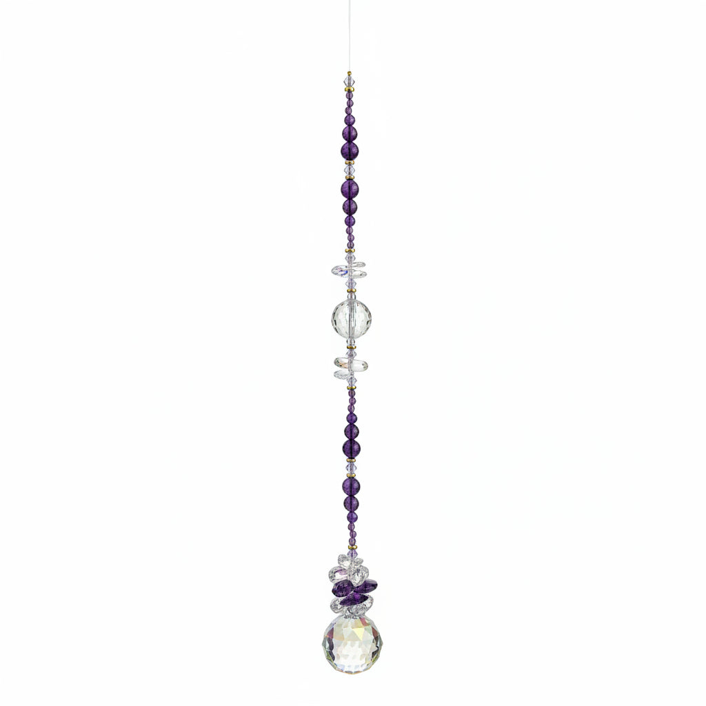 A beaded suncatcher or wall hanging with dark purple and clear faceted beads, a small cross, and a large iridescent crystal pendant, laid flat on a brown wood floor.