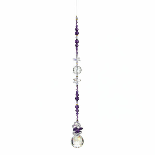 A beaded suncatcher or wall hanging with dark purple and clear faceted beads, a small cross, and a large iridescent crystal pendant, laid flat on a brown wood floor.