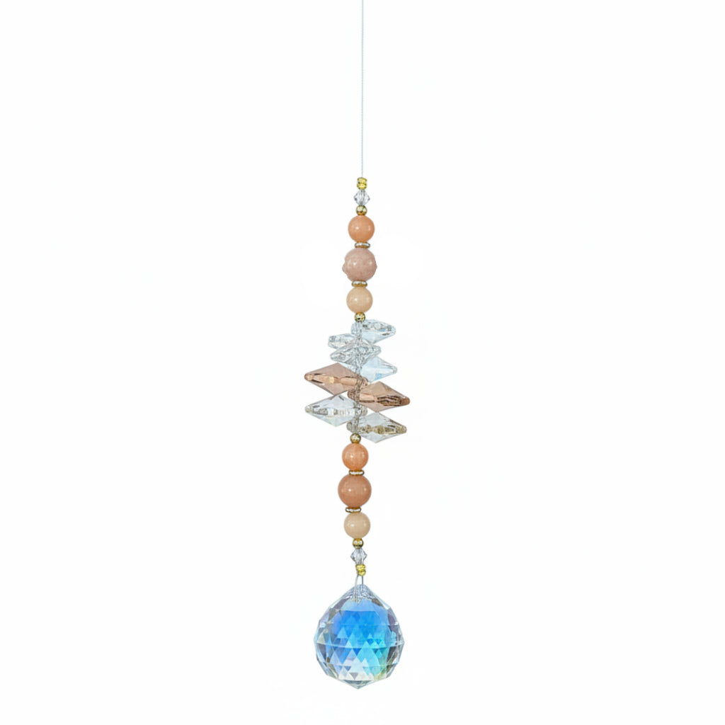 A beaded hanging ornament with a large faceted blue teardrop gemstone at the bottom, smaller peach and cream beads, iridescent crystal pieces, and a small printed tag at the top, photographed on a wood surface.