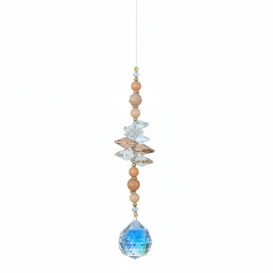 A beaded hanging ornament with a large faceted blue teardrop gemstone at the bottom, smaller peach and cream beads, iridescent crystal pieces, and a small printed tag at the top, photographed on a wood surface.