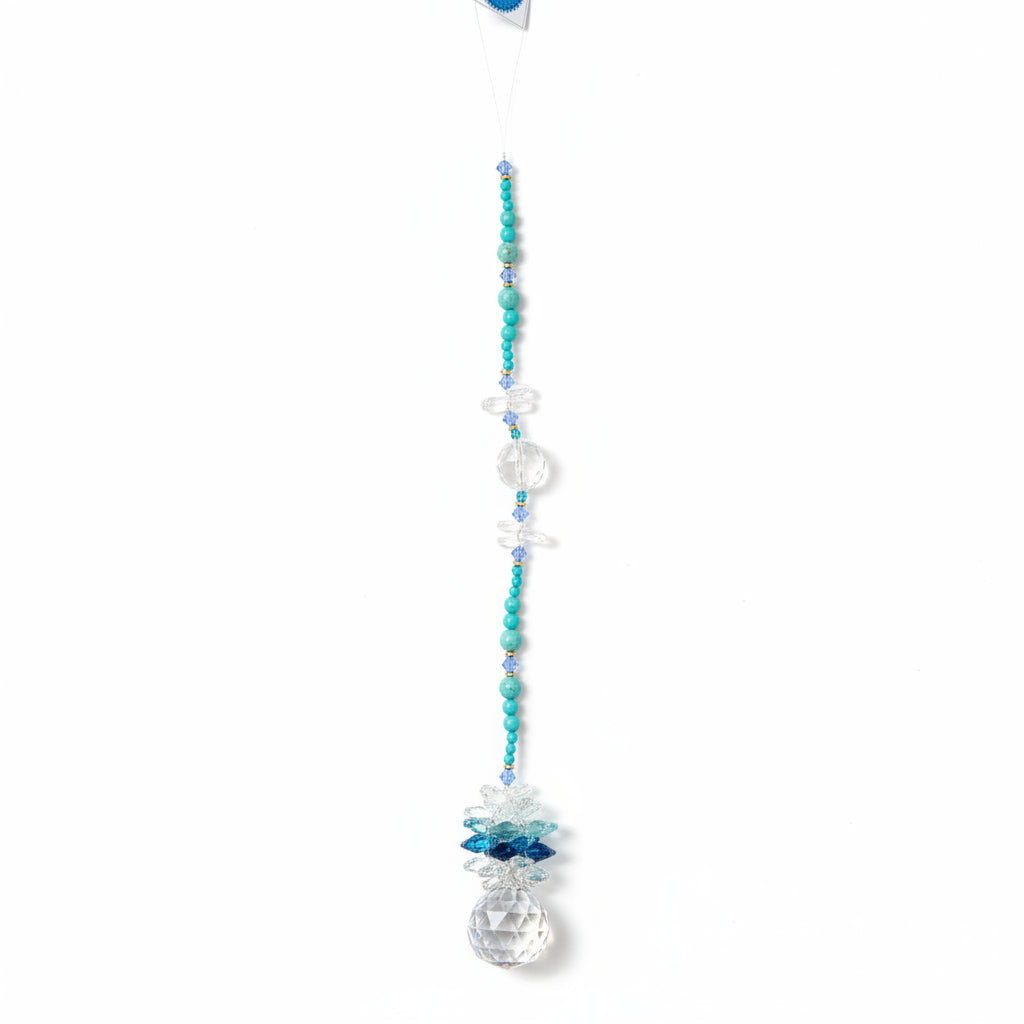 A beaded crystal suncatcher or wall hanging with turquoise blue beads, clear faceted crystal spheres, and a large clear teardrop crystal pendant, laid flat on a brown wood plank floor.