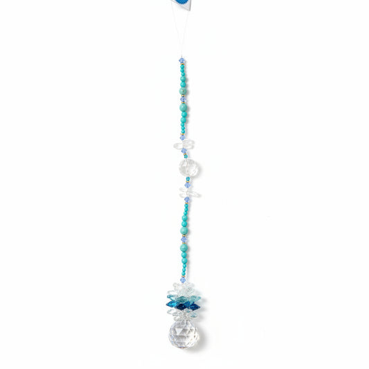 A beaded crystal suncatcher or wall hanging with turquoise blue beads, clear faceted crystal spheres, and a large clear teardrop crystal pendant, laid flat on a brown wood plank floor.
