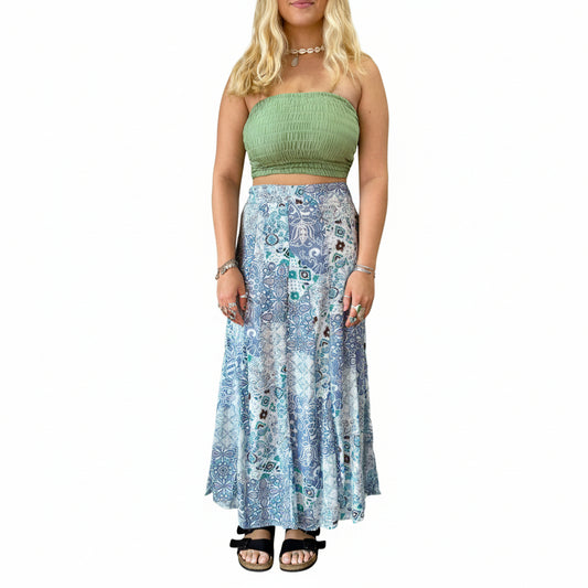 Woman wearing a green top and blue patterned skirt in a room with plants and decor.