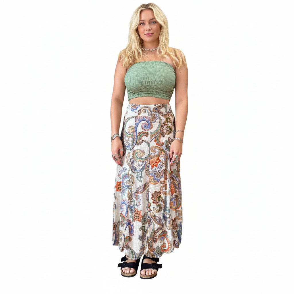 Person wearing a green top and patterned skirt standing in a room with plants and shelves.