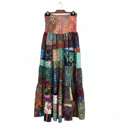 A sleeveless tiered patchwork maxi dress with a smocked bodice, featuring a vibrant multicolor print collage, hanging on a black hanger in a retail store. A hand is visible pointing to the skirt on the left.